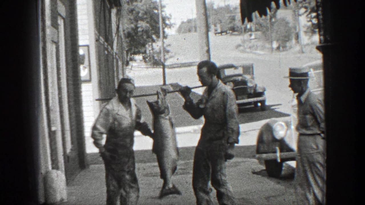 Two men hold up a large fish while another man watches from the sidewalk.