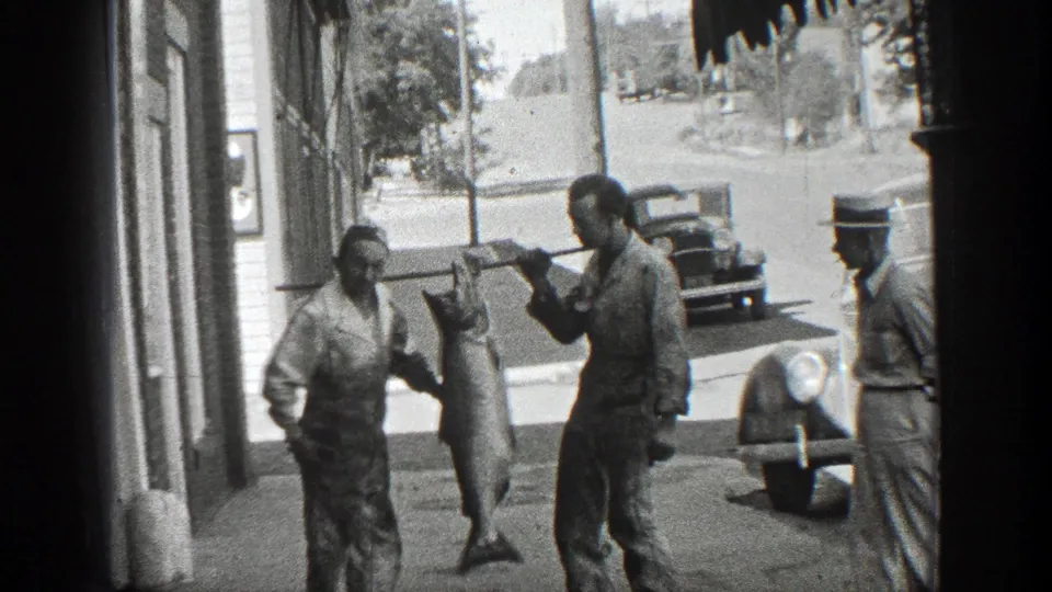 1936 California sidewalk scene with men in period attire showing a large fish near storefronts and parked automobiles.