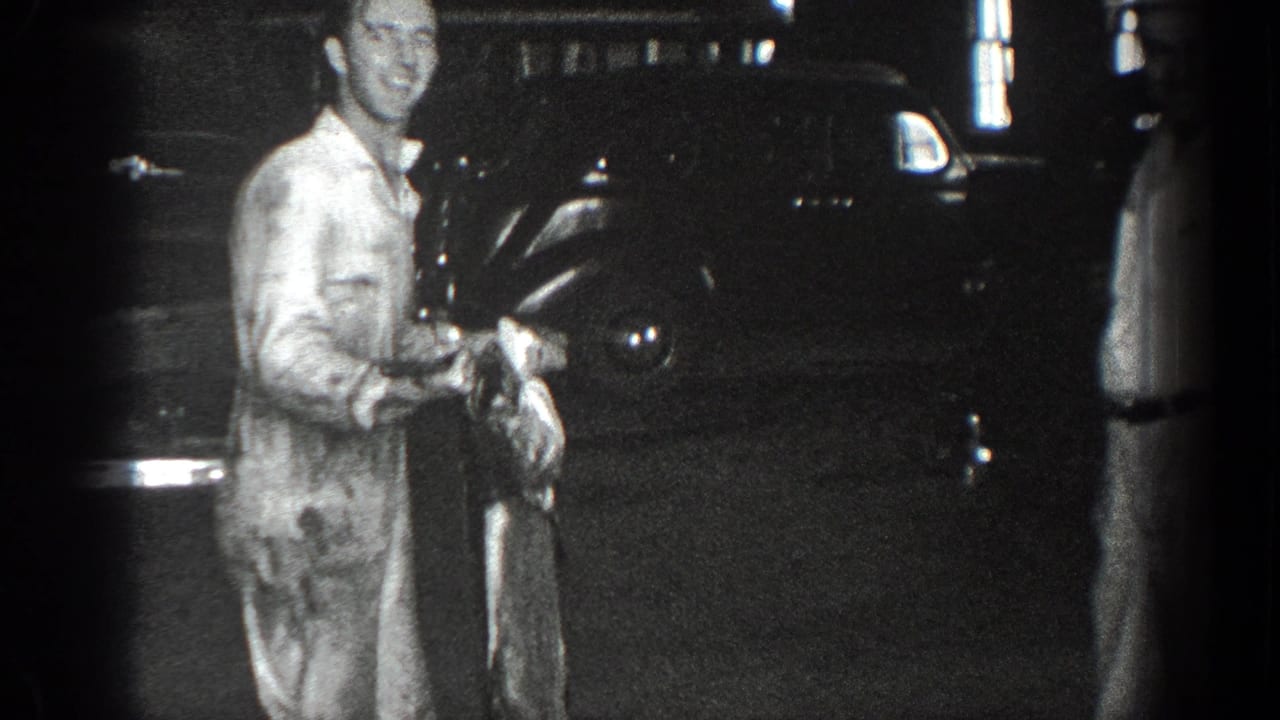 Man in a light jumpsuit stands near a dark automobile at night holding a long object.