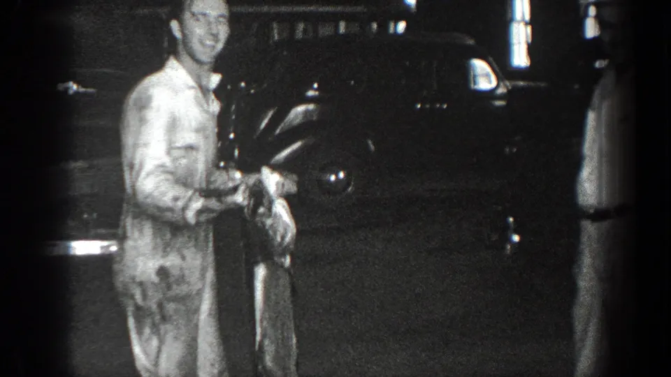 In 1936 California 8mm footage, a man in a light jumpsuit faces the camera near a dark automobile while holding a long grey object.