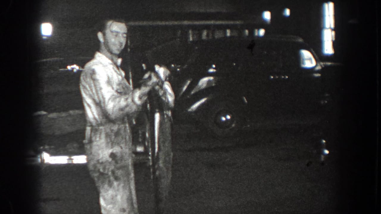 Man in a light jumpsuit holds a long object while standing near a dark car at night.