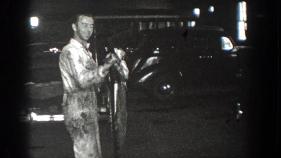 The man in the light jumpsuit turns slightly beside a dark automobile in the nighttime 1936 California home-movie scene.