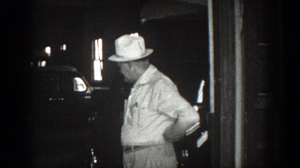 A man wearing a white hat stands in profile beside a dark automobile near a doorway in the 1936 California footage.