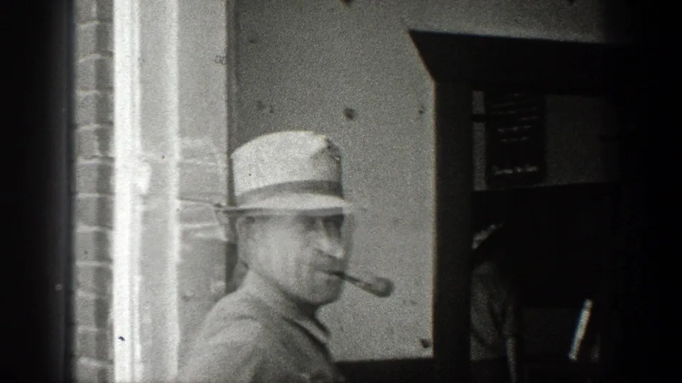 A man in a white hat appears near a building entrance with a pipe in his mouth in the 1936 California home-movie footage.