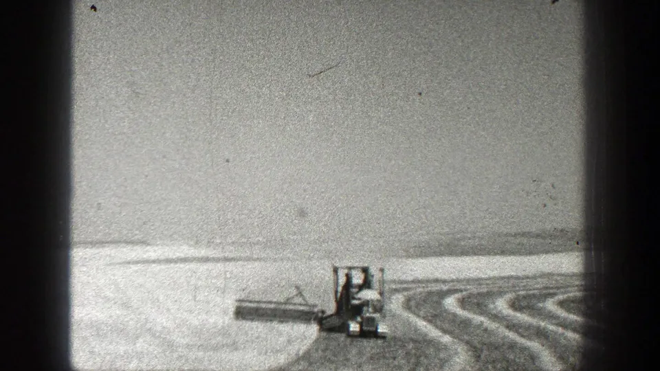 The tractor and attached implement continue through the open field, with the frame showing heavy grain and dark vertical edge shadows.