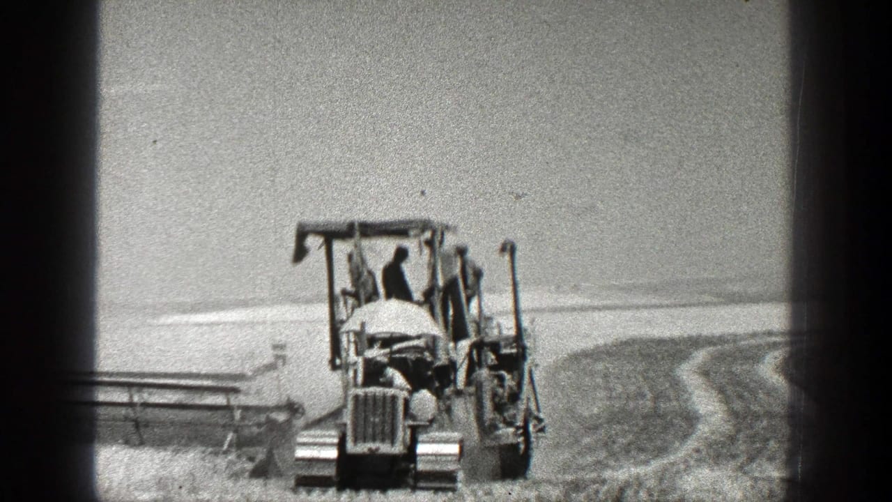 Front view of a tractor and operator in a field with curved plowed rows nearby.