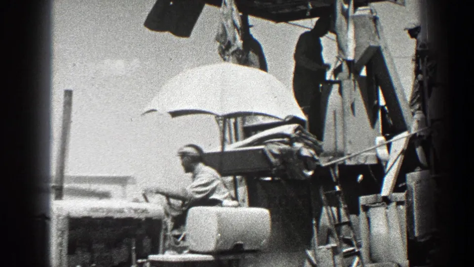 The frame shows a farm worker seated on equipment while nearby laborers stand on the machinery structure in 1936 California footage.