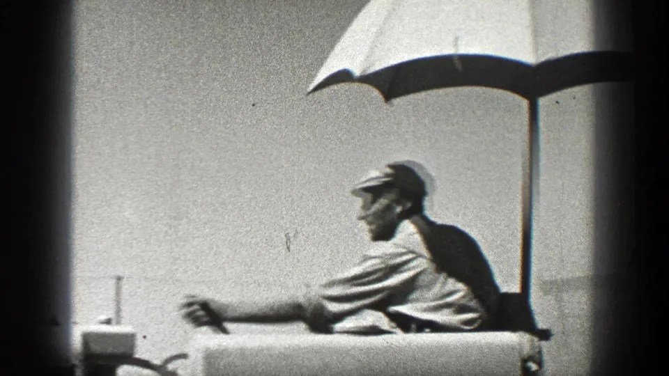 1936 California home-movie frame of a worker in a cap leaning near equipment under a shade umbrella.