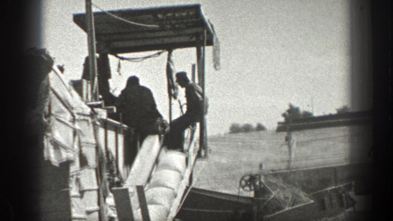 Men ride or work on a large farm machine under a canopy in a field.