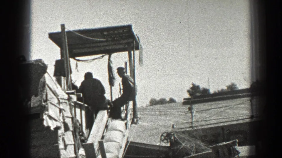 1936 California home-movie frame showing workers on a heavy farm machine, with a large field and distant horizon visible.