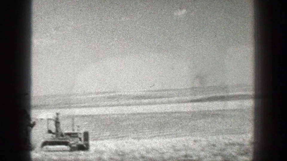 A hazy black-and-white 8mm frame shows the field and partial agricultural equipment as the machine moves through the landscape.