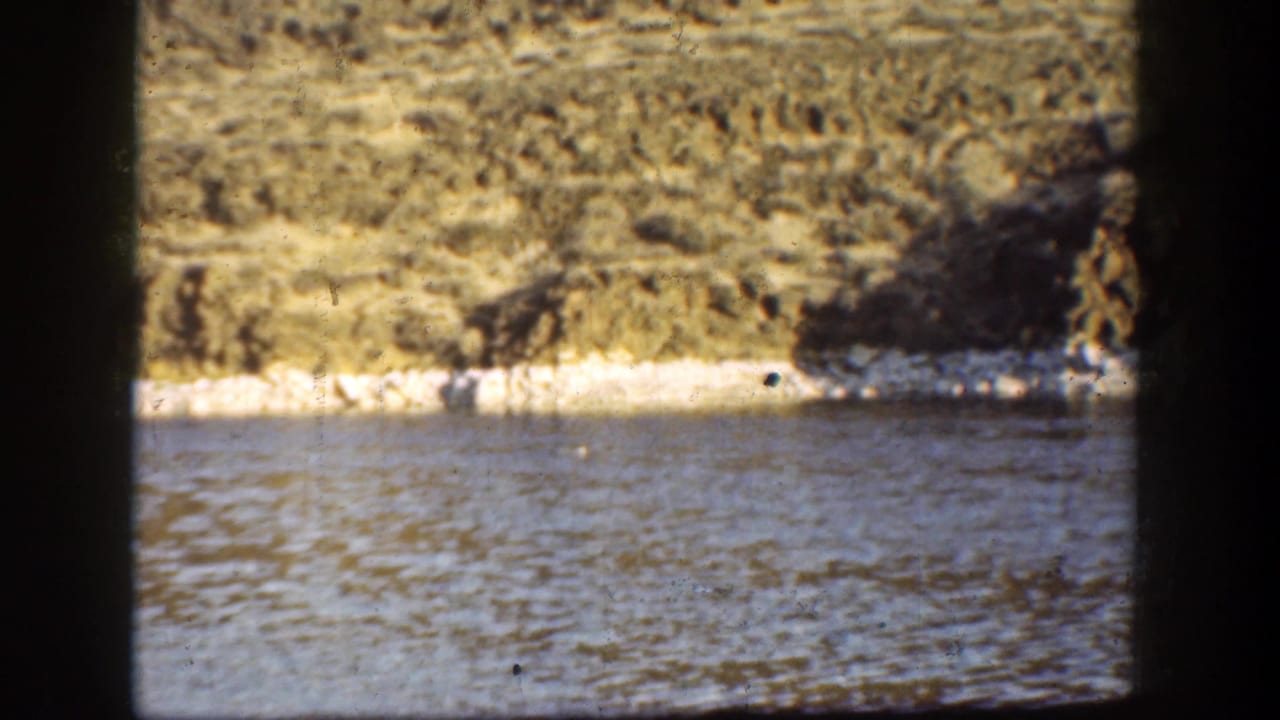 Lake surface and pale shoreline beneath a dry hillside, partly bordered by dark frame edges.