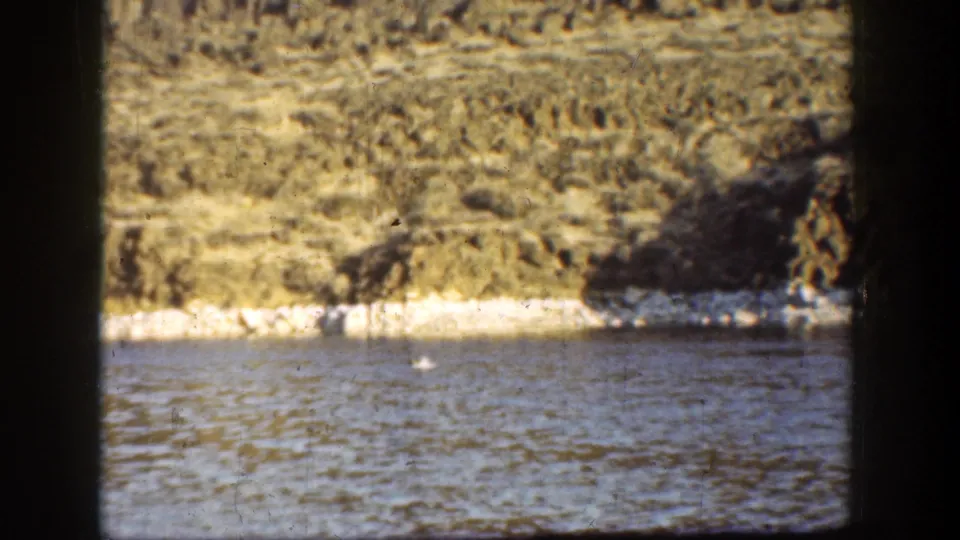 1936 California 8mm frame with a small white object floating near shore against water and a rugged hillside.