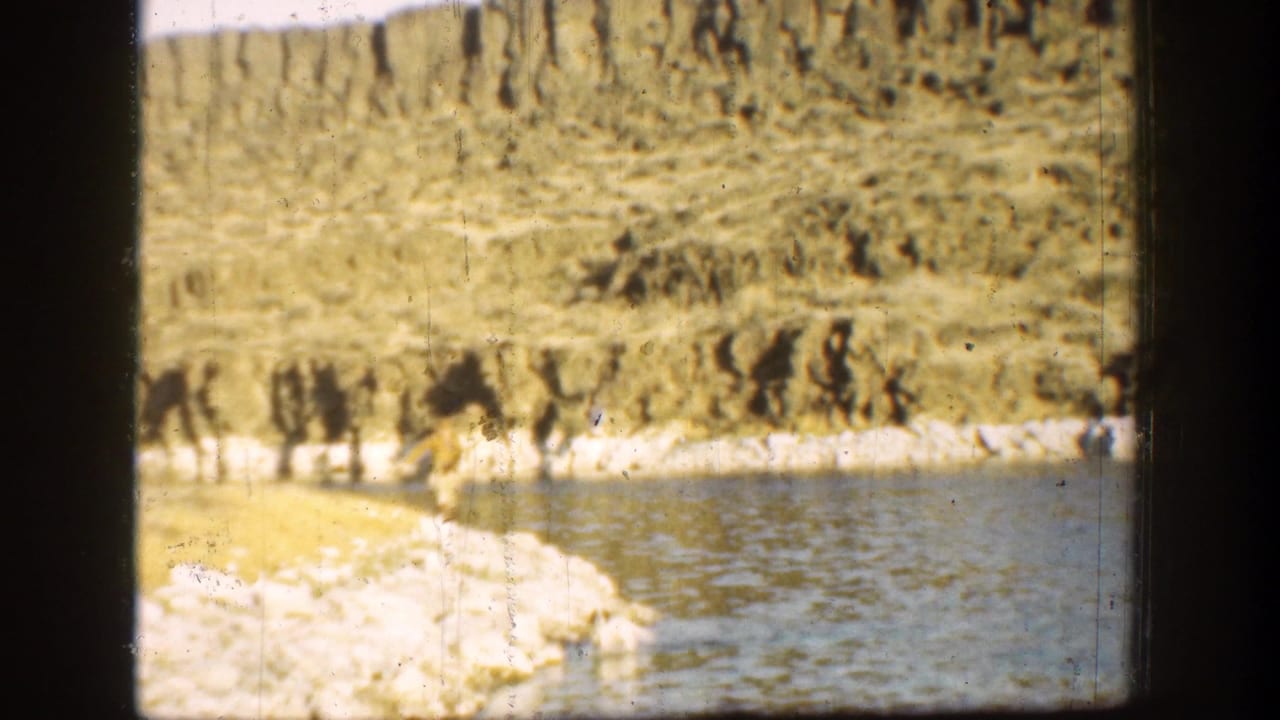 Man stands near a rocky water edge below layered hills in a grainy 8mm frame.