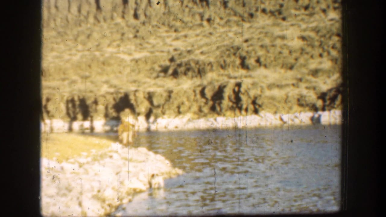 Man walks along the pale rocky bank beside rippled water and hillside terrain.