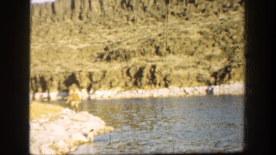 1936 California home-movie frame with a distant figure at the rocky waterline, backed by pale rocks and brushy hillside.