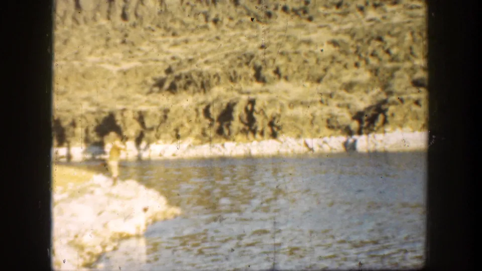 Faded 8mm frame from 1936 California showing a man by the rocky water edge with rippled water and hillside in the background.