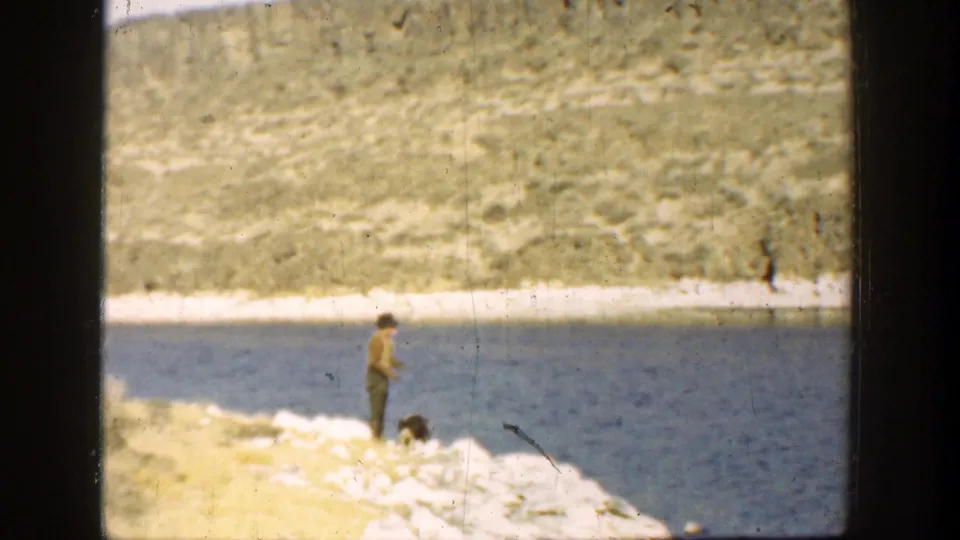 A faded 8mm 1936 California frame shows the man watching as the dog moves low along the rocky lakeshore.