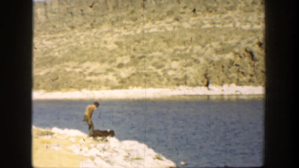 1936 California 8mm frame showing the man leaning toward his dog on the bright lakeshore, with blue water and dry hills behind.