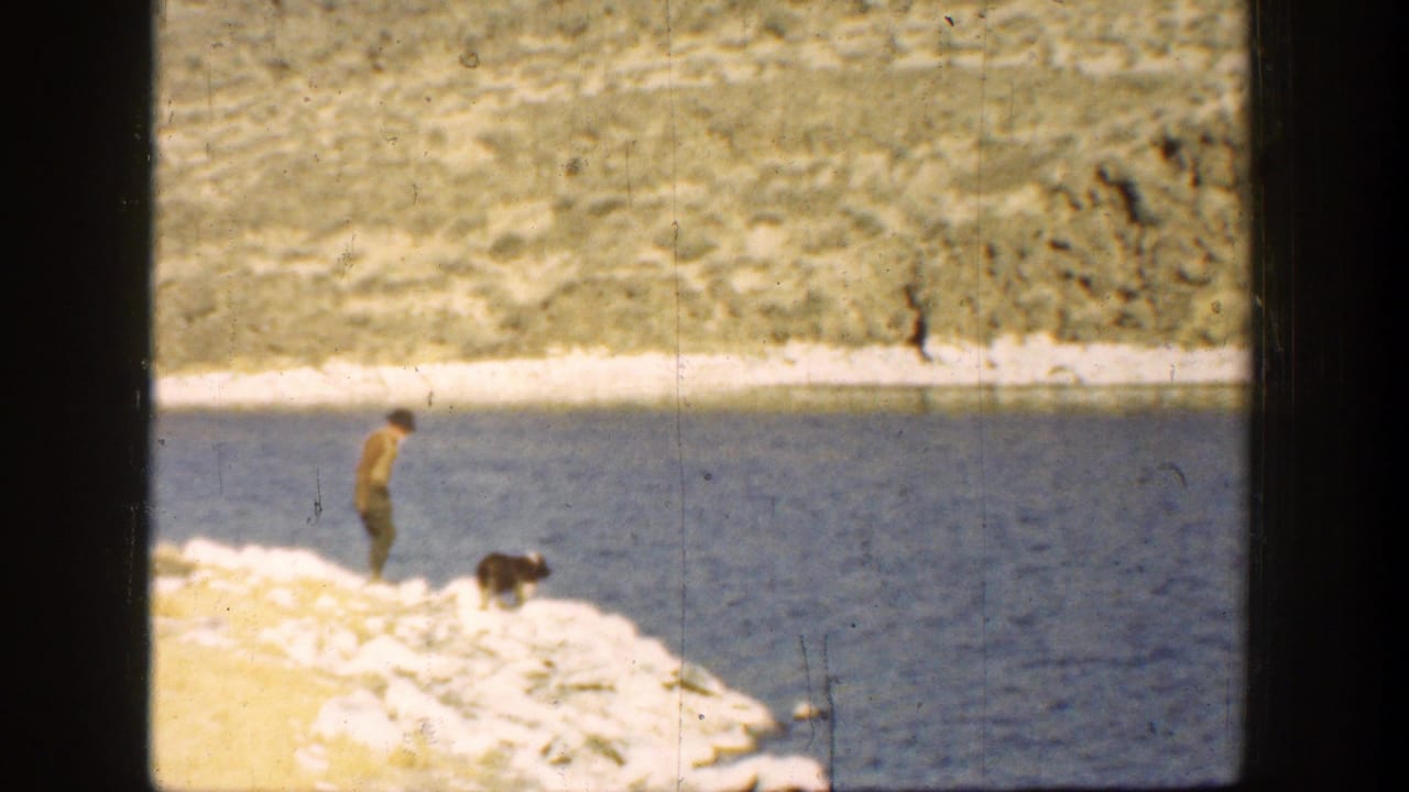 Man and dark dog stand on a pale rocky riverbank beside blue water.