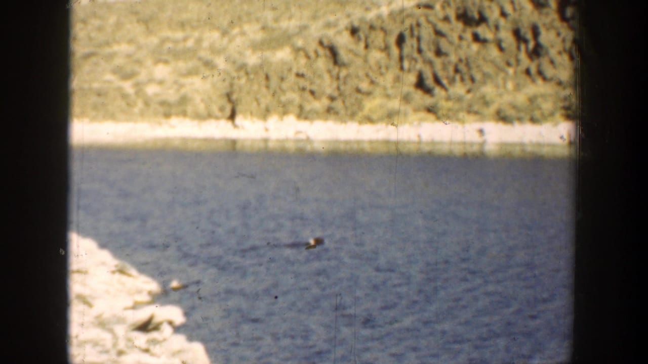 Distant floating object on blue water with rocky shore and hillside behind it.