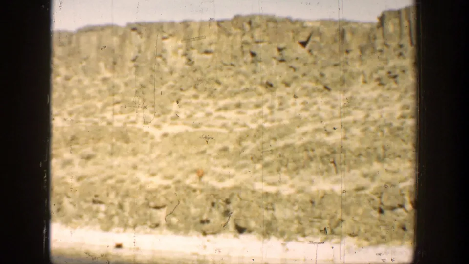 1936 California coastal frame showing dry rocky terrain and distant eroded hills, with part of an adult figure cropped at the right edge.