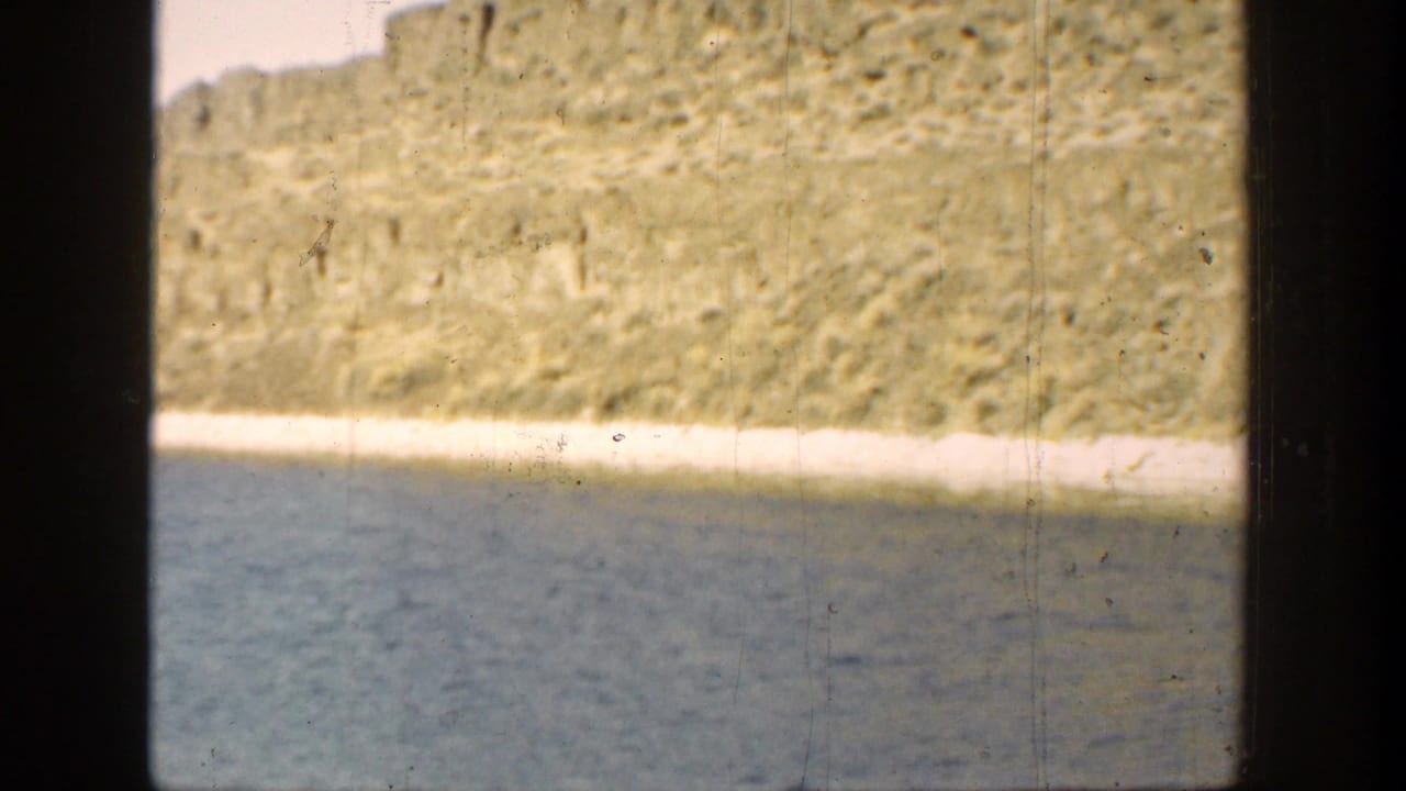1936 California home-movie frame showing calm lake water, a bright shoreline, and a textured cliff face with visible film gate edges.