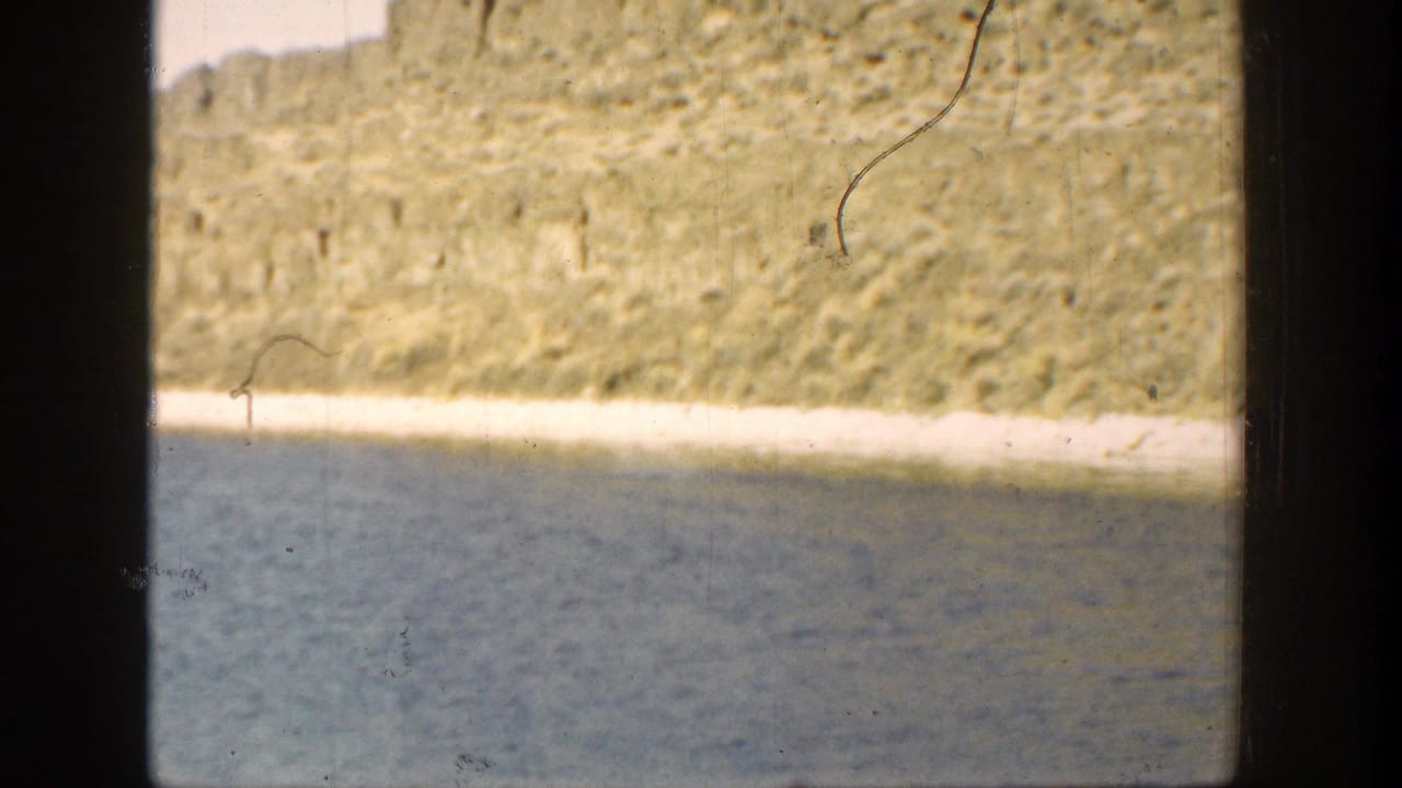 Rocky lakeshore and blue water with a dry hillside behind it, framed by dark film edges.