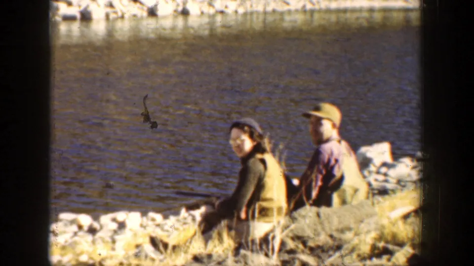 1936 California home-movie frame showing two adults seated on a rocky lakeshore in cool-weather clothing, apparently fishing or resting.