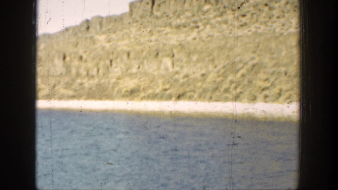 Calm blue water below a pale shoreline and rugged cliff within a dark 8mm frame.
