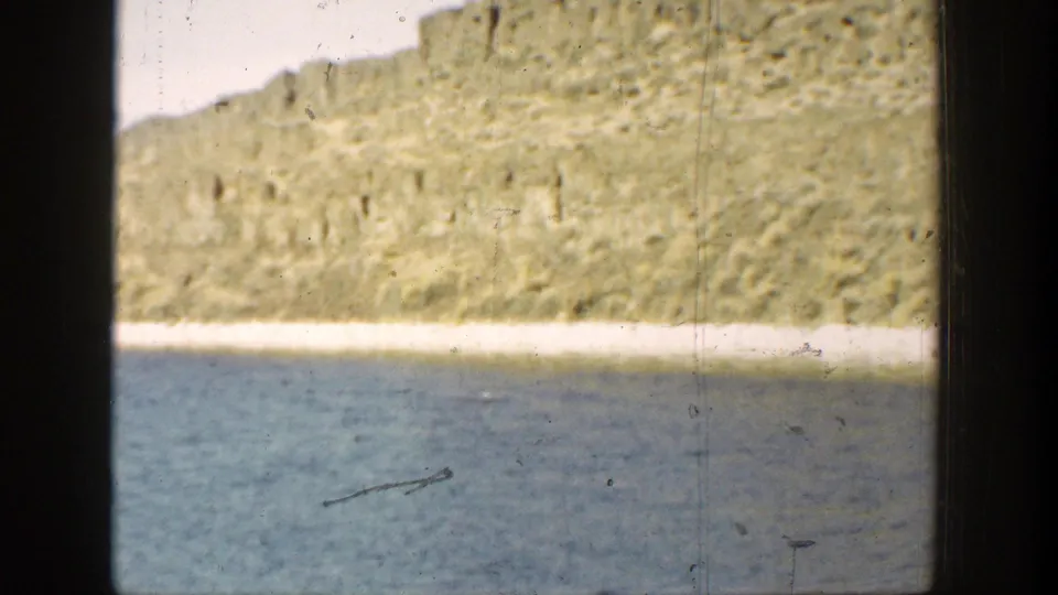 1936 California home-movie frame with calm water, pale shore, rocky cliff, and a small dark linear shape near the lower water.