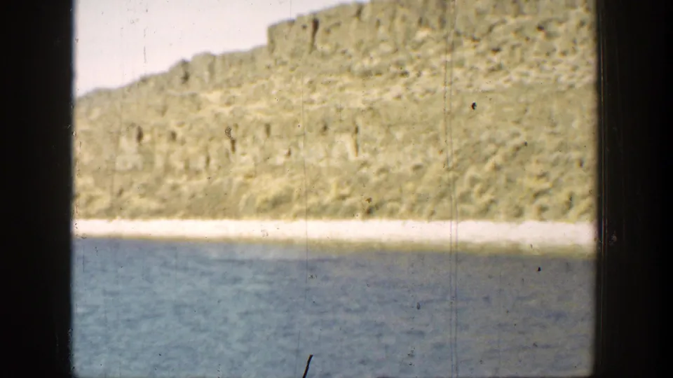 1936 California 8mm frame showing calm blue water, a pale beach line, sunlit rocky terrain, scratches, and a dark frame edge.