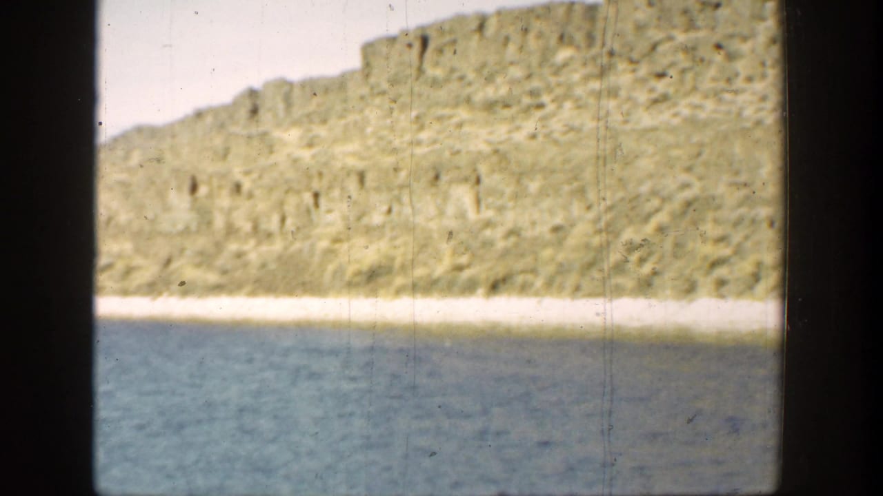 1936 California home-movie frame showing a dry hillside above blue water, with dark 8mm gate edges and visible film wear.