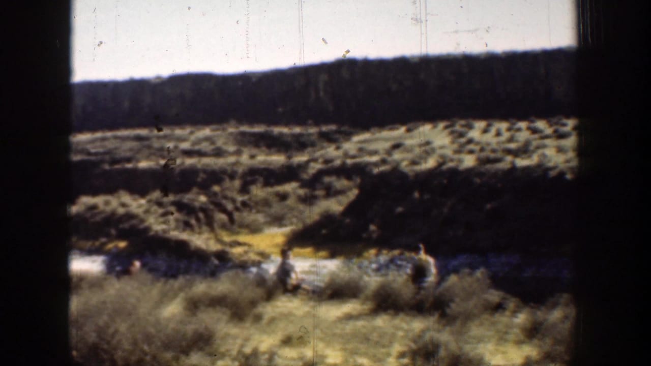 Two men stand near scrub and water below a dark ridge.