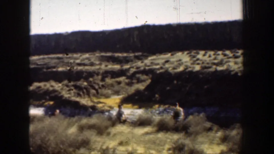 Two men appear near brush and a waterway in the California desert, framed by rugged terrain and a dark film border.
