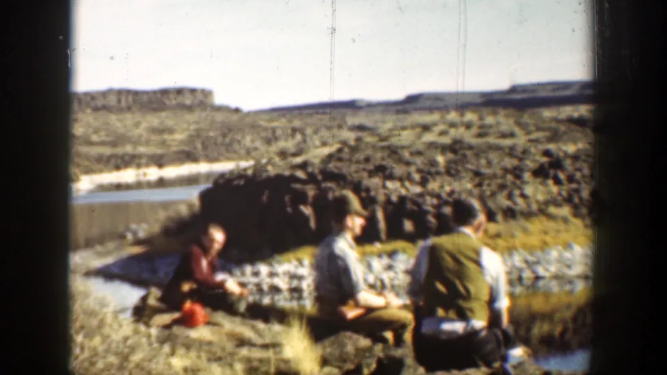 Men in period clothing sit along a rocky riverbank in a 1936 California desert home movie, with hills behind them.