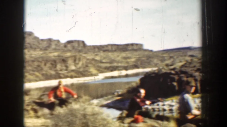 A man in a bright shirt crosses the riverbank while others sit near rocks and water in the California desert setting.