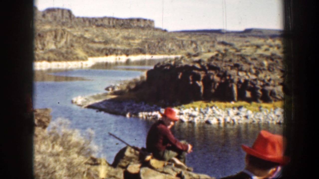 Man in red hat sits on rocks near a river holding a rifle.