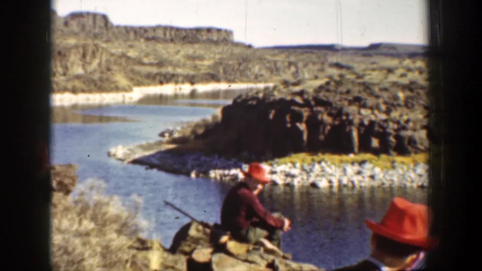 1936 California home-movie frame showing a man in red hat on rocky ground by a river with a rifle nearby.