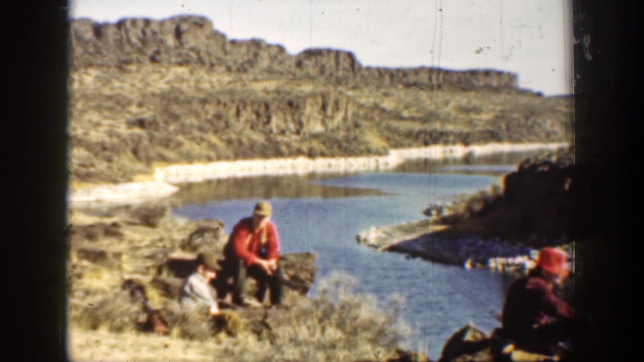 Two men sit on rocks near a river while another red-hatted figure is partly visible.