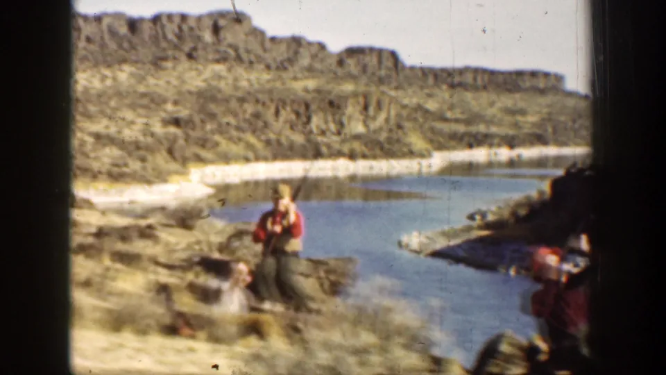 1936 California home-movie frame of a man holding a rifle near a river, with rocky canyon cliffs behind him.