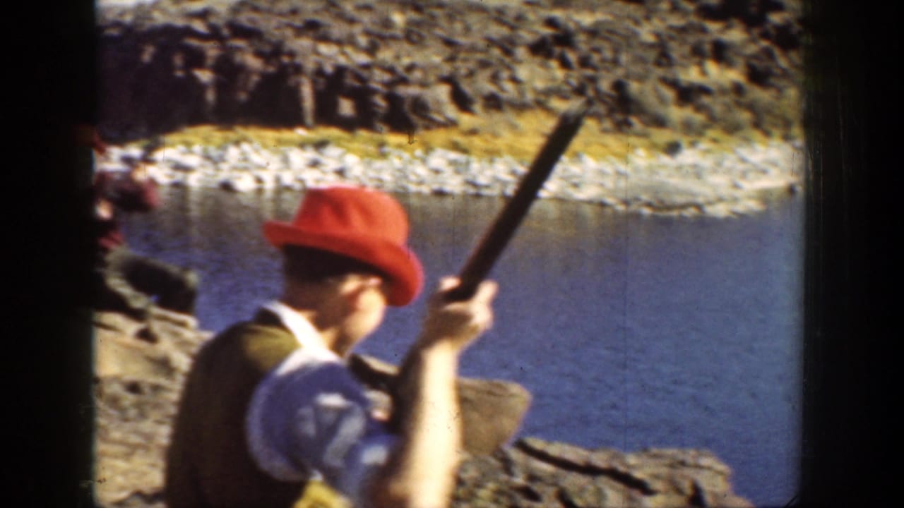 Man in a red hat holds a rifle near a rocky riverbank.