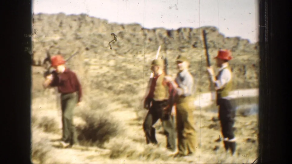 1936 California home-movie frame showing armed hunters in red hats pausing on a brushy path below rocky hills.