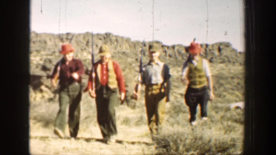 Three men in period outdoor clothing carry rifles near the camera, with a fourth partly visible beside dry brush and rocky terrain.