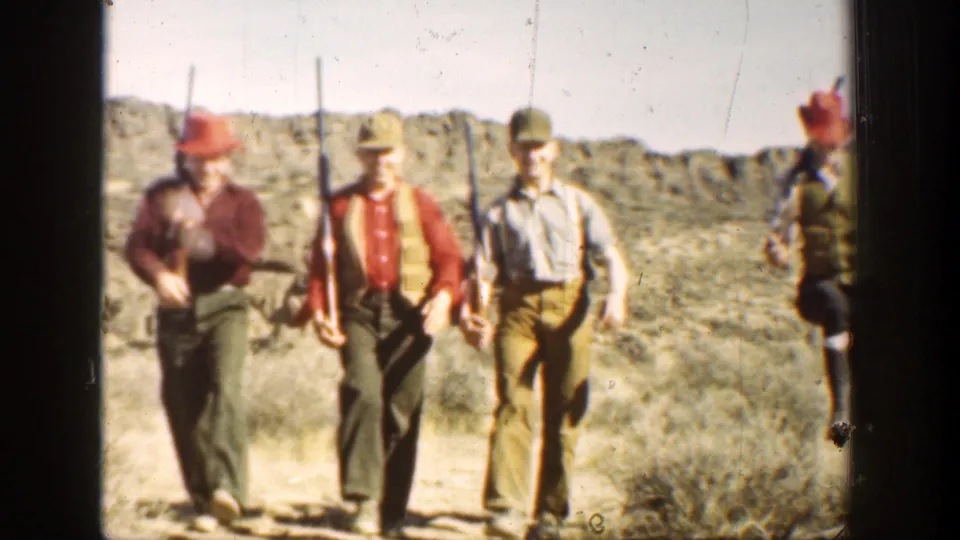 Near the end of the shot, three men with rifles pass close to the camera in a sunlit arid California setting, with one figure partly cropped.