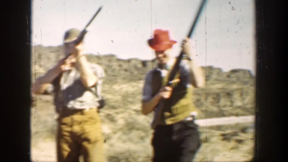 1936 California 8mm frame showing two men in caps and vests carrying long rifles against dry hills.