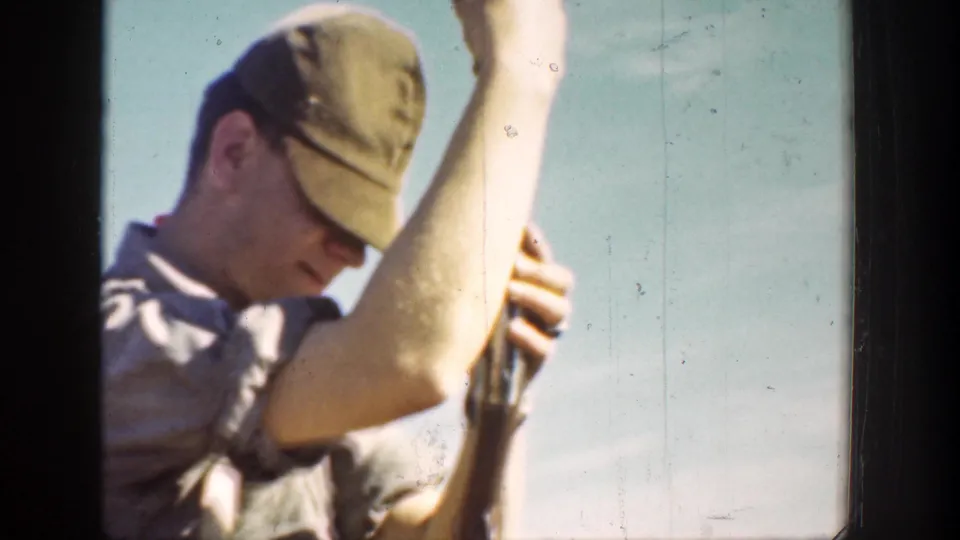 Close 1936 California 8mm frame of a man in a cap holding a rifle upright under a pale sky.
