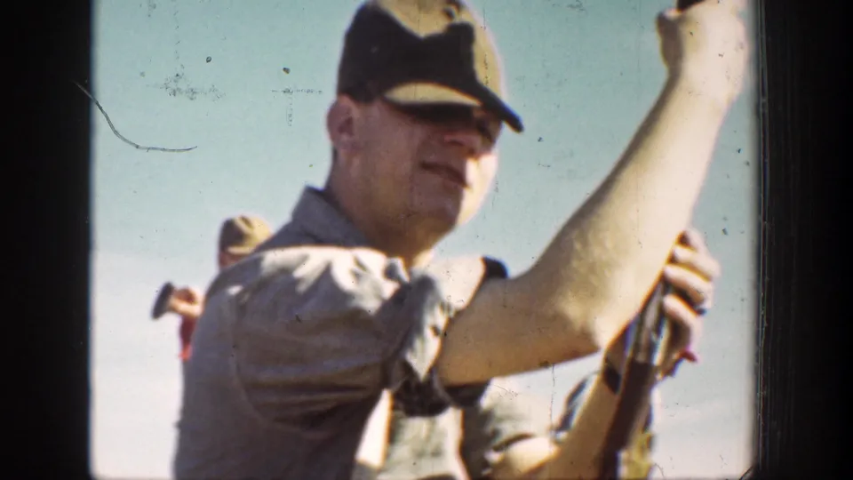 1936 California 8mm frame of a man in a dark cap holding a rifle upright, with another man partly visible behind him.