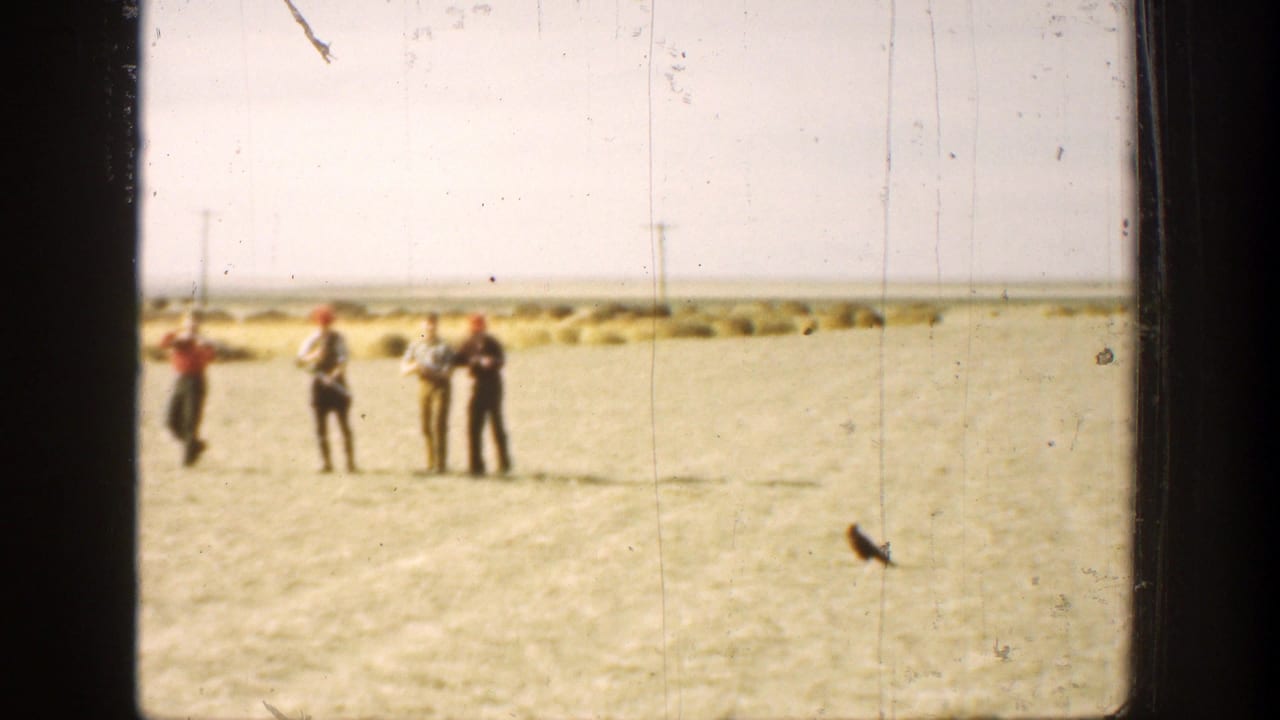 Four men stand in a dry open field, with some holding long guns or gun-like objects.