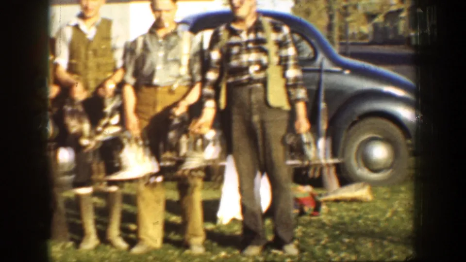 Adult hunters in period clothing stand with ducks near a dark car, with strong sunlight and soft 8mm focus.